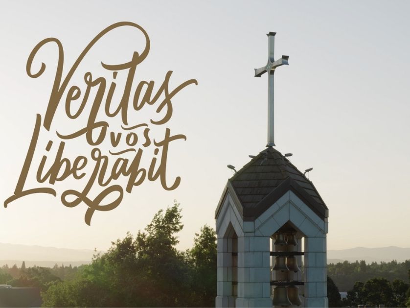 A church bell tower with a cross on top stands beside script text reading Veritas vos Liberabit, which means The truth will set you free. Trees and distant mountains are visible under a clear sky.