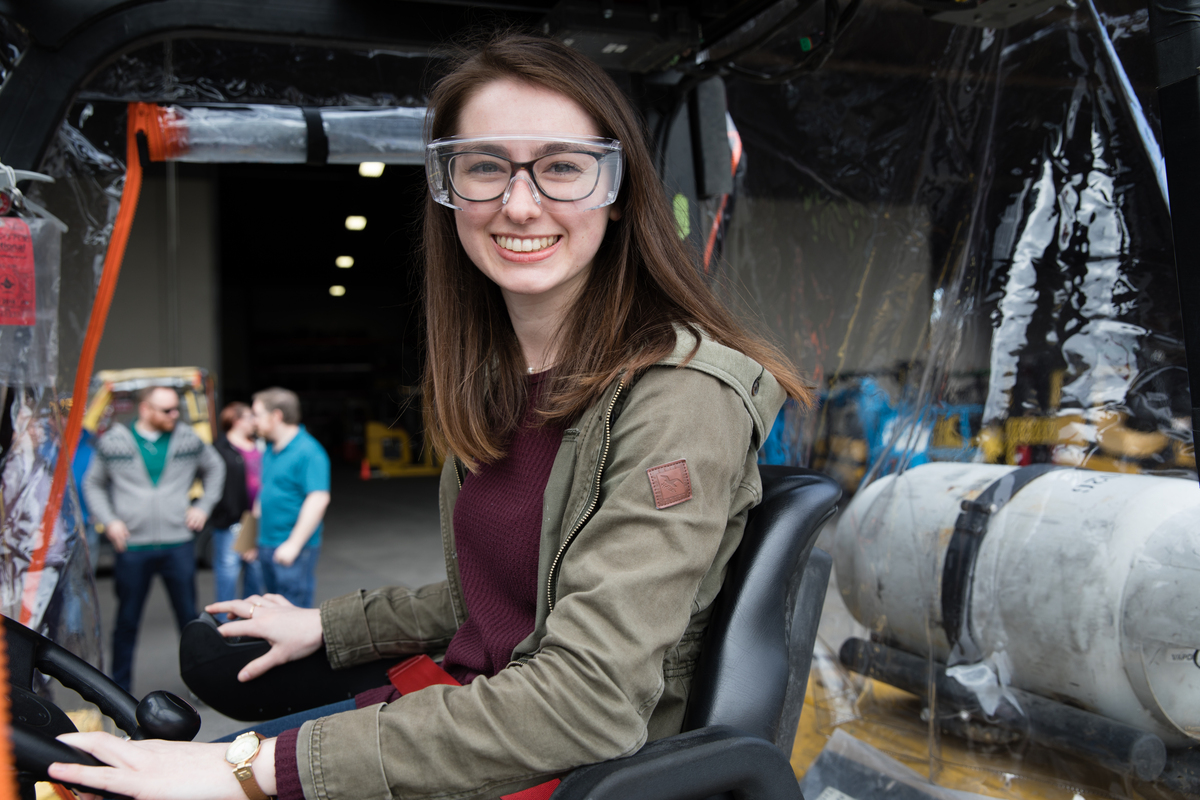 A girl sits on a forklift with safety glasses smiling.