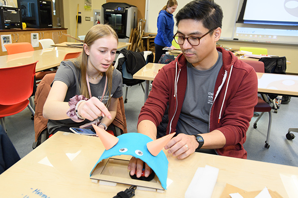 Two students sit at a desk manipulating a paper project