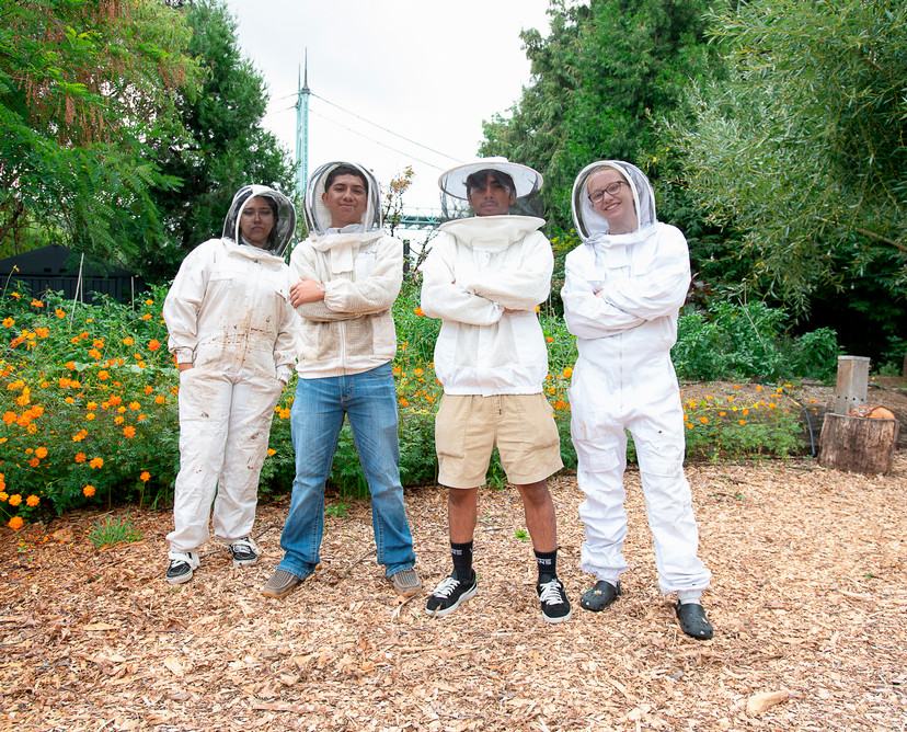 Three students in beekeeper suits stand in front of bee hives in a garden