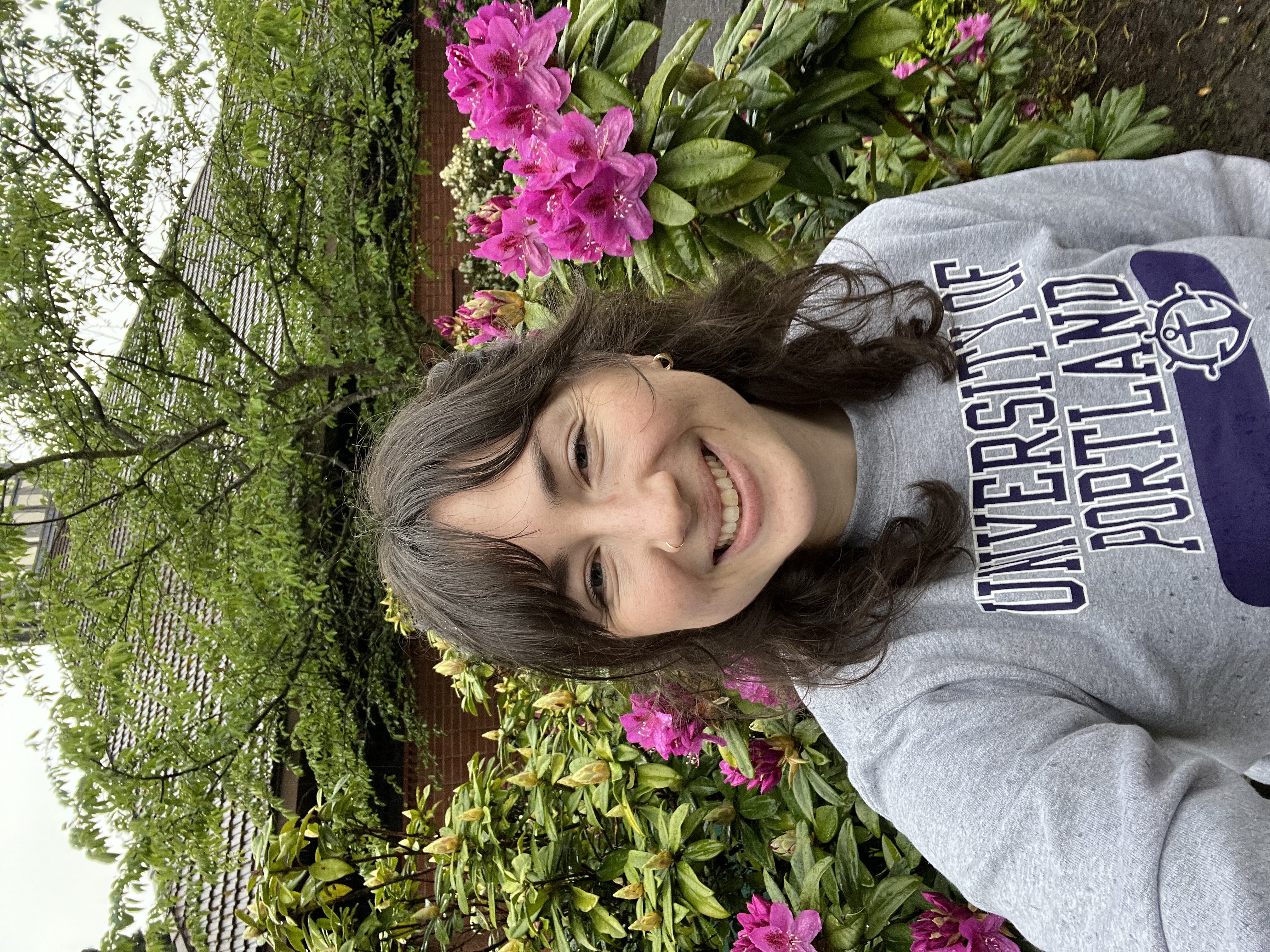 White woman with blue eyes and dark hair smiles at the camera with purple flowers and a brick building in the background
