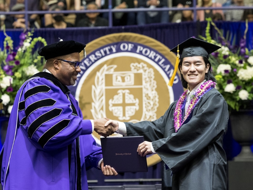 President Kelly shakes hands with a graduate on the dais while handing them a diploma.