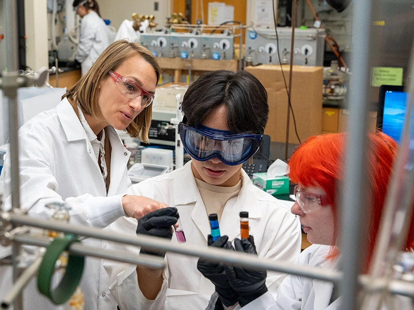 A teacher stands with two students in a lab setting