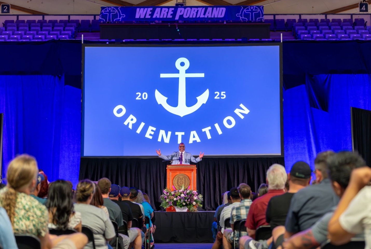 President Kelly with his arms raised during the Welcome address with screen that says 'Orientation' behind him. 