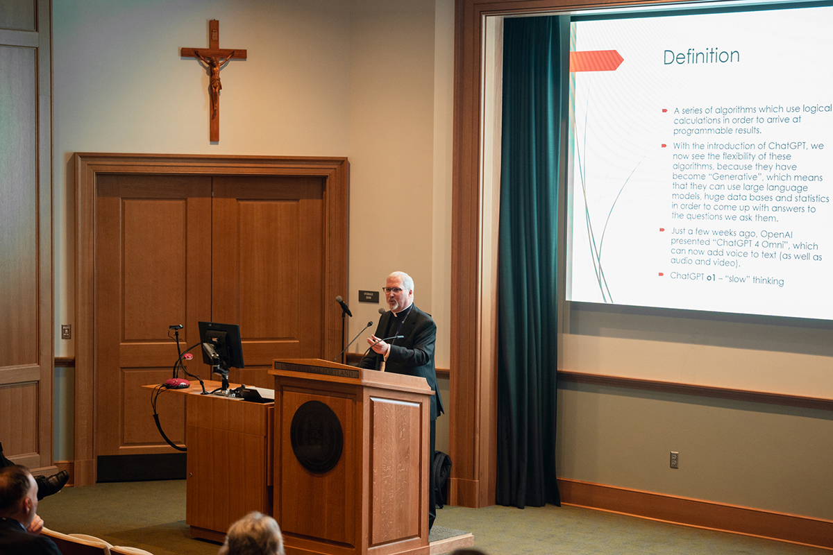 Speaker stands in front of audience with presentation slide in background.