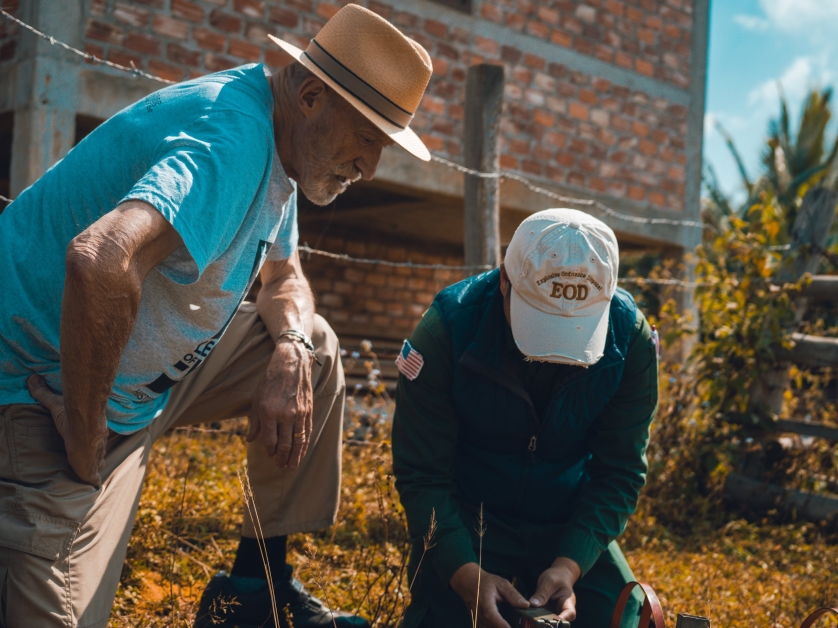 Mike Burton kneels alongside a volunteer crouched over a detonator at they prepare to remotely detonate an unexploded ordinance in Laos.