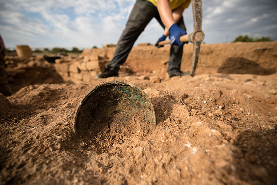A ground level view of an archeological dig site with an ancient bowl front and center, while a person stands poised to dig with a pick axe in the background.