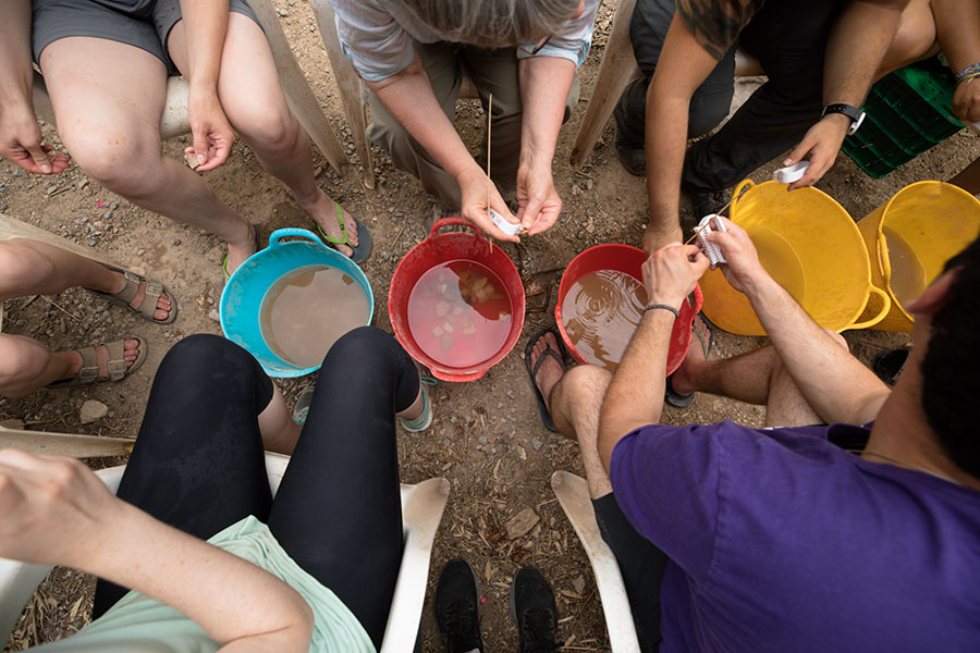 An overhead view of a row of colorful buckets filled with murky water, lined by student hands on either side washing pot sherds