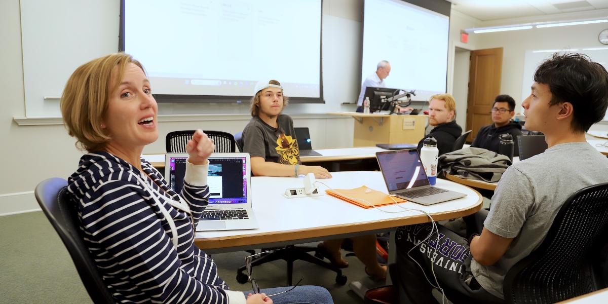 Students and teachers sit in a modern classroom with two projector screens extended over a white board.