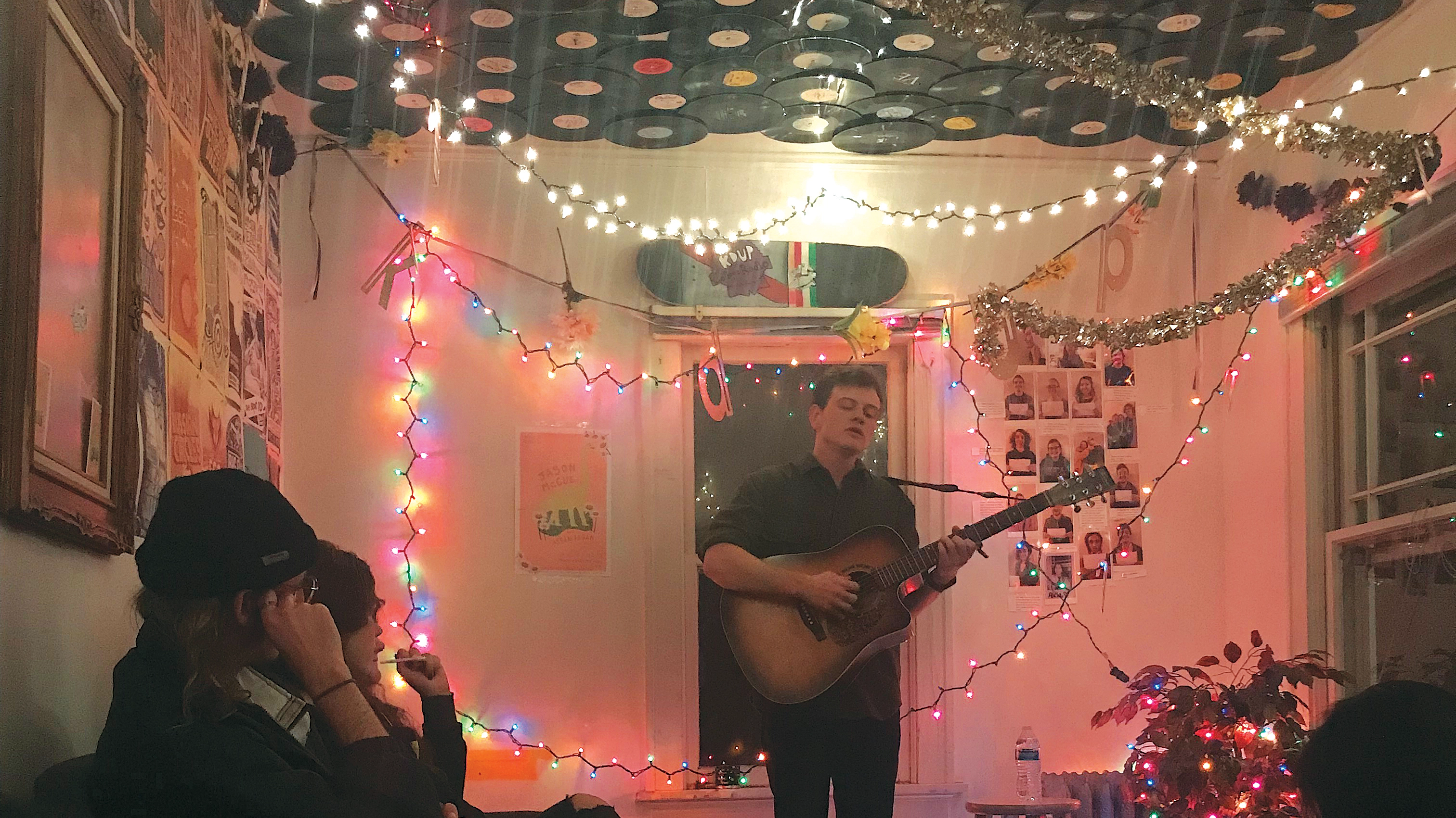 A performer with a guitar stands in front of a microphone in an informal setting at a small gathering