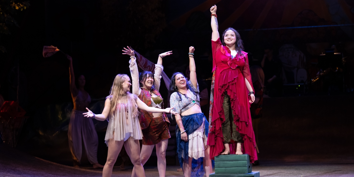 Maddie Tran stands on several crates with her right arm raised high at the center of the image, surrounded by three female actresses who also have their hands outstretched in a scene from the Portland Center Stage production of the musical Hair.