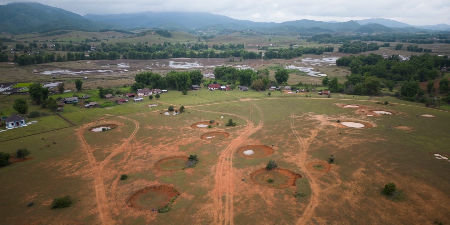 An aerial image of a farm field with rolling green hills in the distance shrouded in mist. The field is dotted with large craters from previous explosions.
