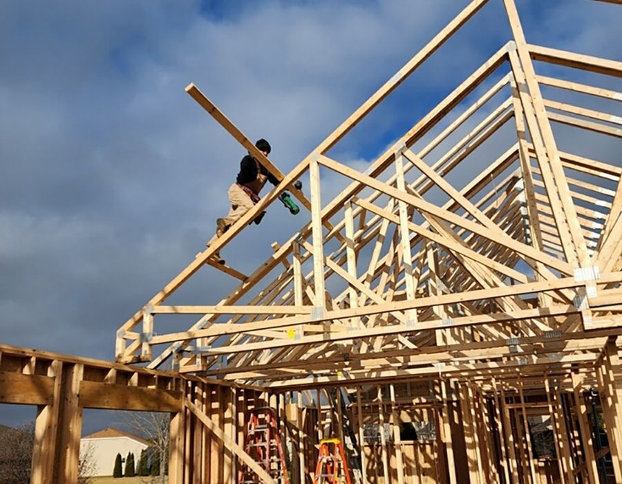 Person working on a wooden house framework with ladders below.