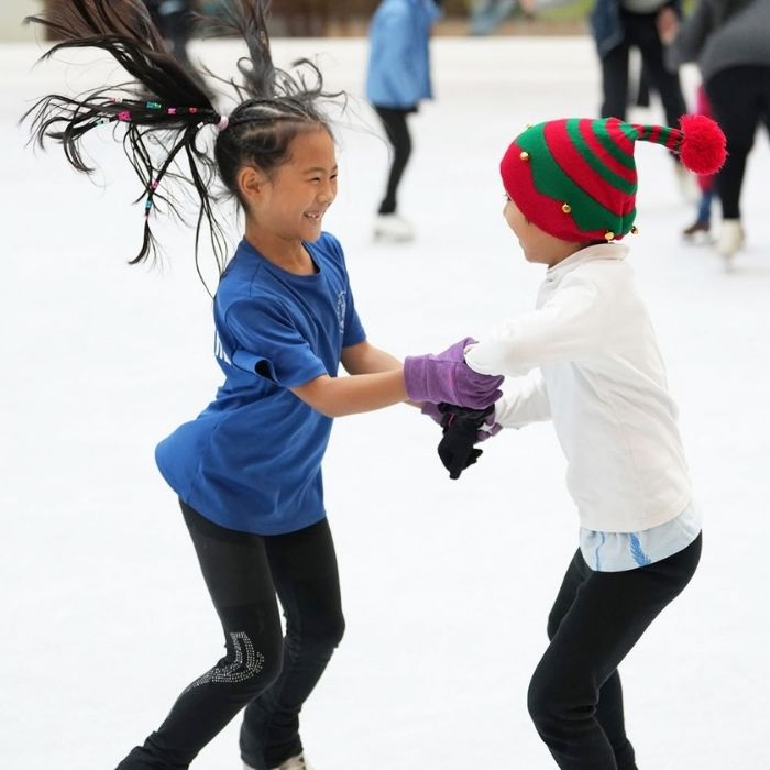 Two child figure skaters face each other and hold hands as they jump on the ice rink.