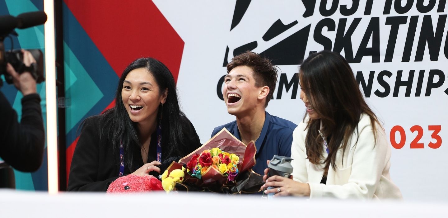 Three people smiling in front of a US Figure Skating Championships backdrop, with one holding a bouquet.