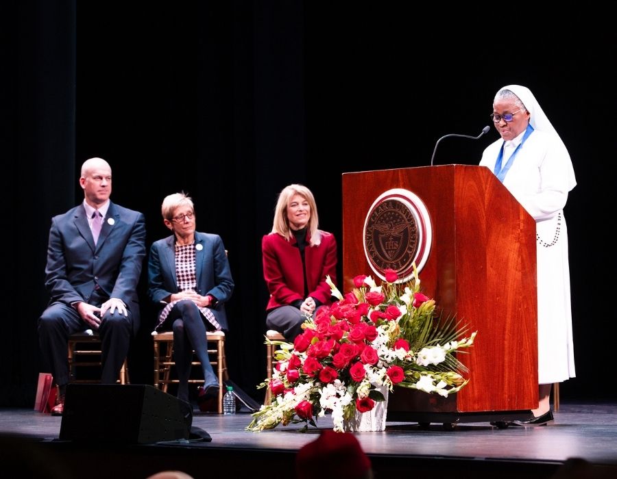 A woman delivers a speech at a podium, surrounded by attentive listeners seated behind her.