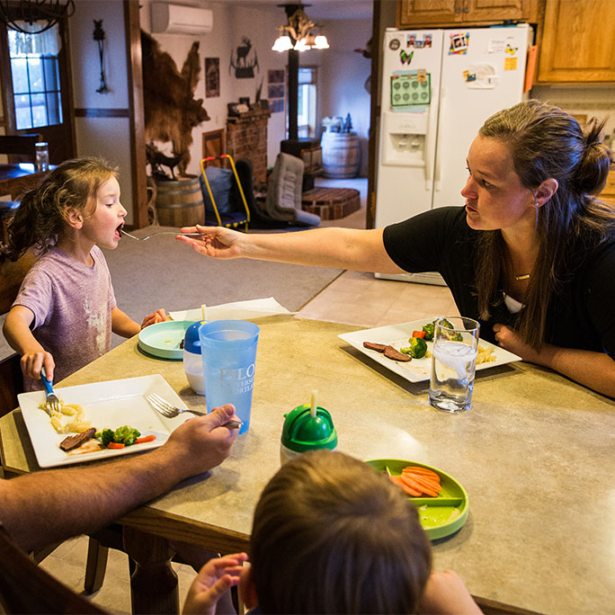 Chelsea sits at a dinner table with two children; she reaches out with a fork to feed her daughter.