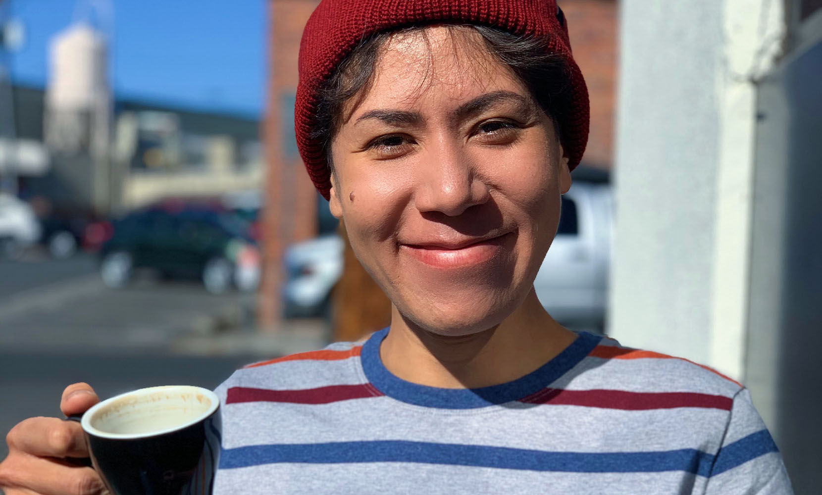 Tara wears a red knit cap, holds up a coffee mug, and smiles