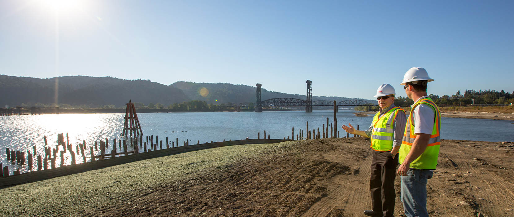 Two men in hard hats stand on revegetated land along the riverbank.