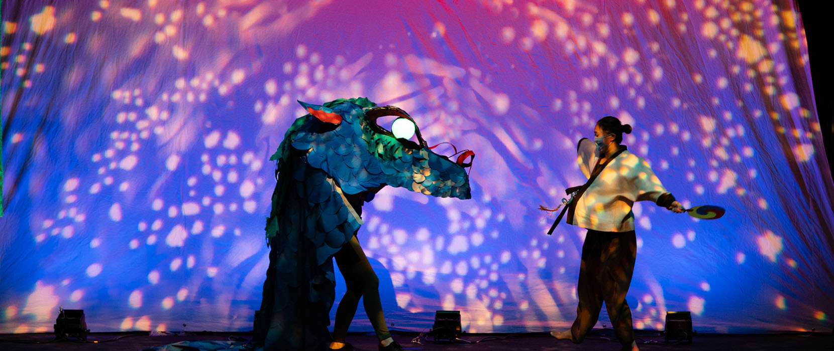 a dancer faces a life-size dragon puppet on a coolly lit stage
