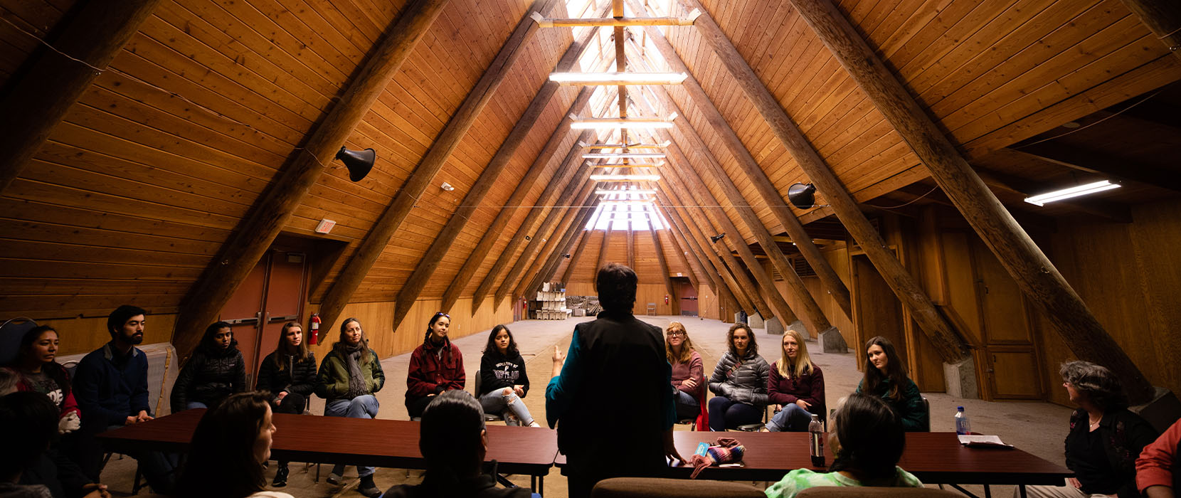 University of Portland students sit in chairs in a Native American longhouse and listen to a speaker