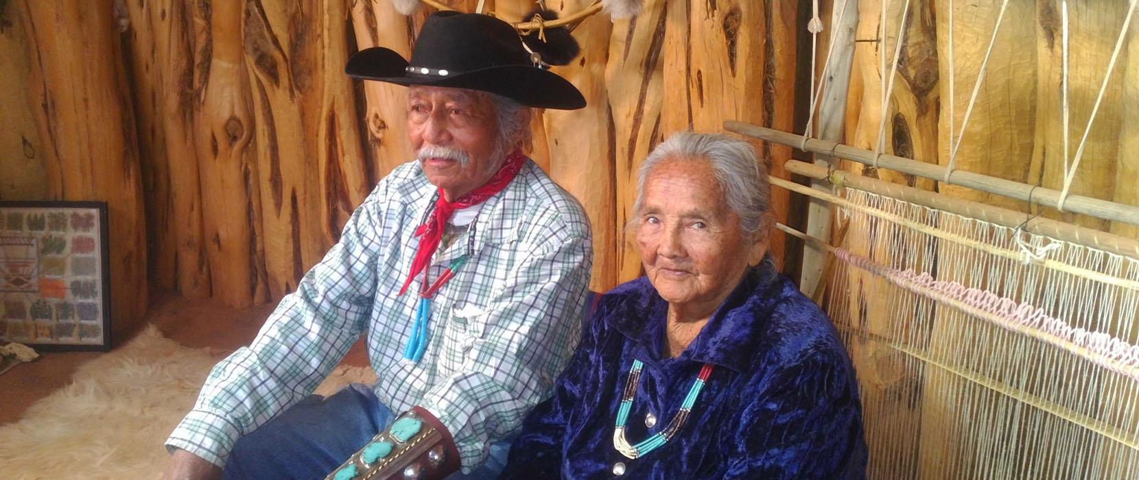 Devonna's grandparents sit in front of a loom. The grandfather wears a black cowboy hat and the grandmother wears a beaded necklace.